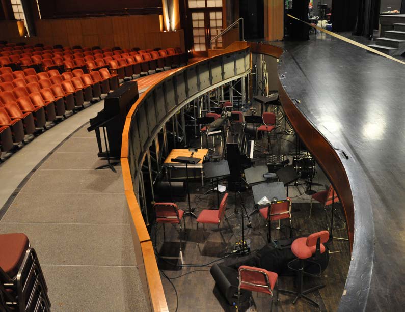 Theater stage with an orchestra pit set up with chairs, music stands, and microphones, viewed beside rows of empty seats.